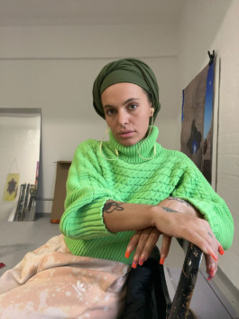 Portrait der Künstlerin Anna Ehrenstein für die Biennale für Freiburg 3, 2025. Sie sitzt seitlich auf einem Stuhl in ihrem Studio und trägt ein grünes Oberteil.