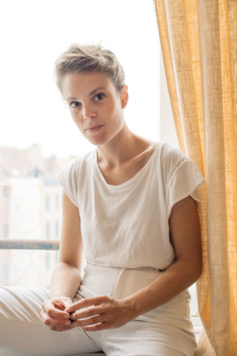 Portrait der Künstlerin Caroline Mesquita für die Biennale für Freiburg 3, 2025. Sie sitzt auf einer Fensterbank mit beigem Vorhang und heller Kleidung.