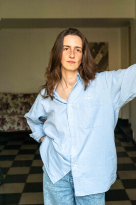 Portrait der Künstlerin Cosima zu Knyphausen für die Biennale für Freiburg 3, 2025. Sie trägt ein hellblaues Hemd und lehnt am Fensterrahmen.