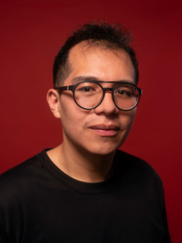 Portrait des Künstlers Ervin Alacron Lam für die Biennale für Freiburg 3, 2025. Er sitzt vor einem roten Hintergrund.