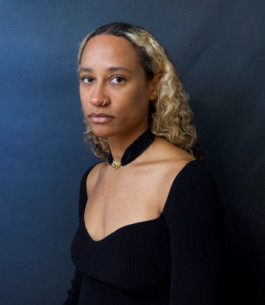 Portrait der Künstlerin Eve Tango für die Biennale für Freiburg 3, 2025. Sie sitzt vor einem dunkelblauen Hintergrund. 