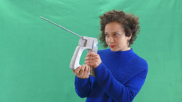 Portrait der Künstlerin Ghita Skali für die Biennale für Freiburg 3, 2025. Sie steht vor einer grünen Wand und hält ein Gerät in der Hand. 