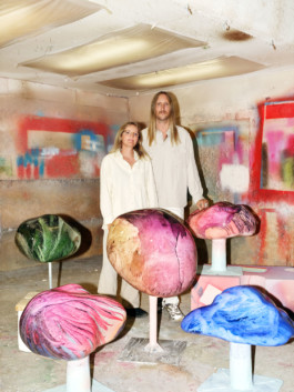 Portrait der Künstler*innen Inka und Niclas für die Biennale für Freiburg 3, 2025. Beide stehen zwischen übergroßen bunten Pilzen und tragen weiße Kleidung.