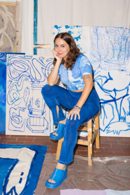 Portrait der Künstlerin Irene Fernandez Arcas für die Biennale für Freiburg 3, 2025. Sie sitzt auf einem Stuhl, vor ihren blauen Malereien.