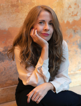 Portrait der Künstlerin Irene de Andres für die Biennale für Freiburg 3, 2025. Sie sitzt vor einer rötlichen Mauer und stützt ihren Kopf mit der Hand auf.