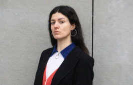 Portrait der Künstlerin Karla Zipfel für die Biennale für Freiburg 3, 2025. Sie steht vor einem grauen Hintergrund und trägt ein weiß, blau, rotes Oberteil.