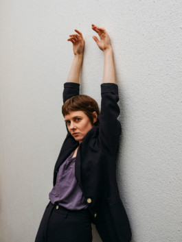 Portrait der Künstlerin Marie Popall für die Biennale für Freiburg 3, 2025. Sie lehnt an einer weißen Wand und streckt die Arme nach oben.