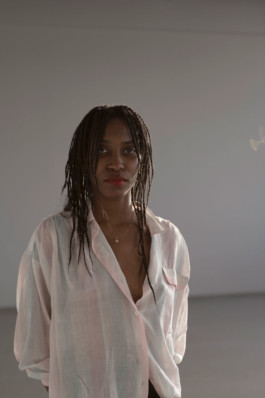Portrait der Künstlerin Monika Emmanuelle Kazi für die Biennale für Freiburg 3, 2025. Sie steht vor einer hellgrauen Wand in einem hellen Hemd. 