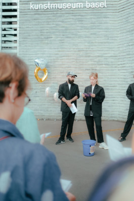 Portrait des Kunstkollektivs Unofficial Hiking Society für die Biennale für Freiburg 3, 2025. Die zwei Personen stehen gemeinsam vor dem Kunstmuseum Basel und lesen etwas vor.