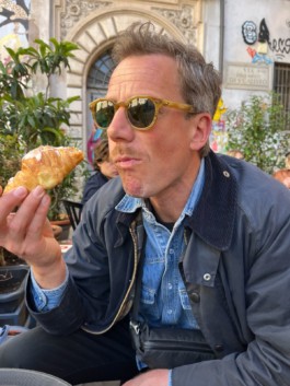 Portrait des Künstlers Florian Thate für die Biennale für Freiburg 3, 2025. Er sitzt mit einer Sonnenbrille in einem Cafe und isst ein Croissant.